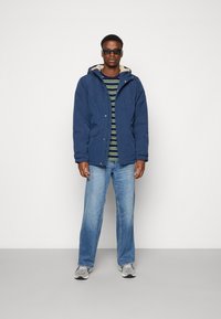 Mørkeblå parka med foret hette, båret over en grønn og hvit stripete skjorte; kombinert med lyseblå jeans og grå sneakers.