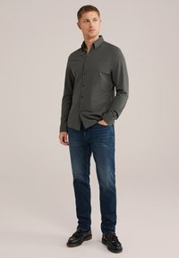 Camisa de botón de manga larga verde oscuro con cuello, combinada con vaqueros azules y zapatos negros. Tejido suave con detalles sutiles.