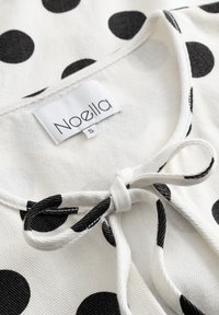 Bílá látka s velkými černými puntíky, na které je u výstřihu uvázaná mašle a malá štítek se značkou velikosti S.