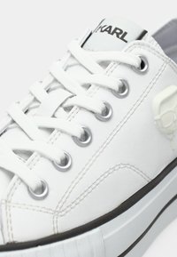 Λευκός δερμάτινος αθλητικός παπούτσι με στρογγυλή μύτη, ασημένιες τρύπες για το κορδόνι και λευκά κορδόνια. Διαθέτει μαύρο λογότυπο στη γλώσσα και υφασμένη λεπτομέρεια στο πλάι.