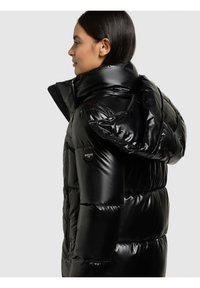 Melnā pūkainā jaka ar spīdīgu virsmu. Tai ir augsts apkaklis, pārizmēra kapuce un logotipa detaļa kreisajā pusē. Aizsargājošs izolēts dizains.