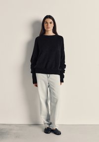 Črn oversized pulover z rebrastimi manšetami, v kombinaciji s svetlo modrimi naravnost krojenimi kavbojkami in črnimi pletenimi sandali. Minimalistična ozadja.
