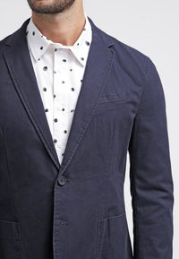 Mörkblå kavaj i bomull med enkel knappstängning, kantskurna slag och bröstficka, lager på en vit skjorta med svarta prickar.