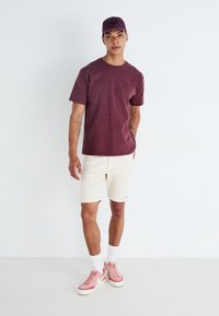 Burgundy puuvillapaita, beige farkkuhousut ja vaaleanpunaiset lenkkarit. Logolla varustettu lippalakki viimeistelee asun. Yksinkertainen, rento muotoilu neutraaleilla väreillä.