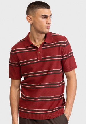 Muž v červenom polokošeli s krátkym rukávom s horizontálnymi čiernymi a bielymi pruhmi, ktorý sa pozerá doprava na jednoduchom pozadí.