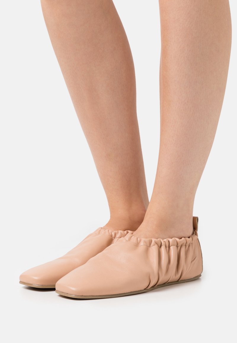 Jambes portant des chaussures légères beiges claires à enfiler, avec des côtés élastiqués froncés et des semelles plates, sur fond blanc.