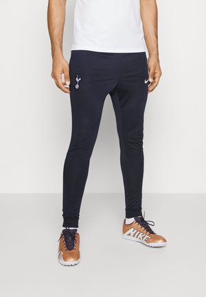 Mann trägt marineblaue Slim-Fit-Sporthose mit Tottenham Hotspur und Nike Logos, kombiniert mit bronzefarbenen Nike Sportschuhen.