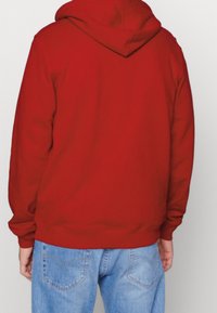Personne portant un sweat à capuche rouge uni et un jean bleu clair, vue de dos sur un fond neutre.