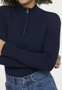 Navy gerippter Rollkragenpullover mit einem halben Reißverschlusskragen und langen Ärmeln, kombiniert mit hoch taillierten blauen Jeans aus Denim mit einem Metallknopf.