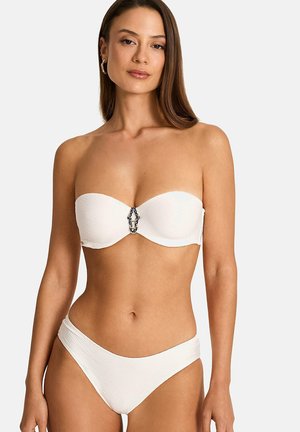 Femme portant un bikini blanc sans bretelles avec une boucle décorative sur le haut, debout devant un fond clair uni.
