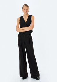 Zwarte vest met v-hals en knoopsluiting, gecombineerd met hoge, wijde zwarte broek. Model heeft het haar sleek naar achteren gestyled.