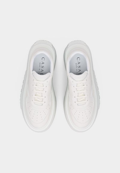 Paire de baskets en cuir blanc avec lacets blancs, zone des orteils perforée et semelles épaisses en caoutchouc transparent, vues de dessus sur fond blanc.