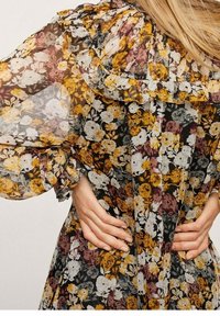 Ohut kukallinen mekko, jossa on keltaisia, valkoisia ja purppuran värisiä kukkia. Mekossa on röyhelökaulus ja puffihihat, joissa on elastiset hihansuut. Kädet lanteilla.