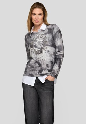 Woman wearing a gray patterned sweater over a white collared shirt and dark jeans, with one hand in pocket, standing against light background.