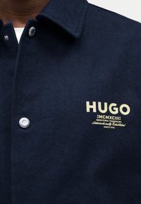 Nærbillede af navy jakke med sølvfarvede trykknapper og beige "HUGO MCMXCIII" logo trykt på venstre bryst.