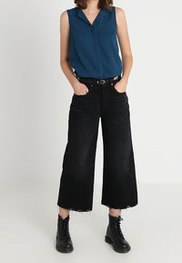 Femme portant un haut sans manches en teal foncé, un jean large cropped noir avec des ourlets bruts, une ceinture noire et des bottes à lacets noires, les mains dans les poches.