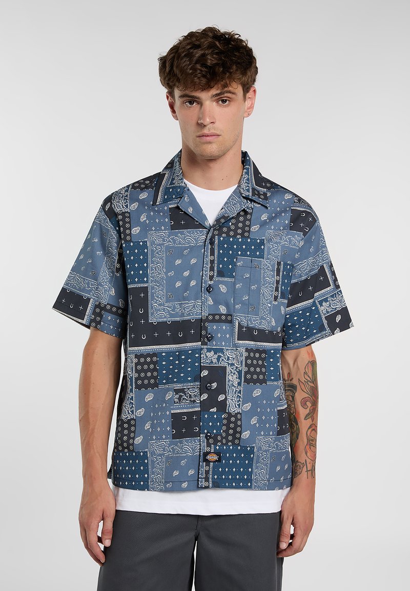 Camicia a maniche corte con bottoni in tonalità di blu, caratterizzata da un design patchwork con motivi e texture vari. Include una tasca sul petto e un colletto piegato.