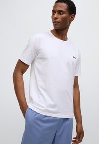 T-shirt bianca a maniche corte in cotone, con scollo rotondo e piccolo logo nero sul petto a sinistra. Abbinata a pantaloncini blu chiaro.