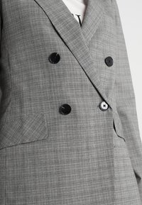 Veste croisée à carreaux gris avec un tissu texturé, des boutons noirs et une poche avant unique.