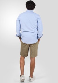 Camisa de botones de color azul claro, pantalones cortos color caqui y zapatillas blancas. La camisa tiene mangas largas, un dobladillo redondeado y detalles de costura visibles.