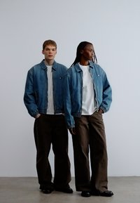 Dvaja modely majú na sebe zhodné svetlomodré rifľové bundy cez biele tričká, v kombinácii s voľnými hnedými nohavicami a čiernymi topánkami, na neutrálom pozadí.