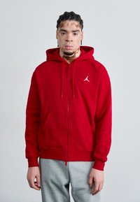 Haut à zip rouge en tissu doux avec des poches kangourou et un petit logo blanc sur la poitrine gauche. Associé à un pantalon de survêtement gris.