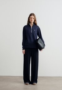 Γυναίκα με ναυτικό μπλε φούτερ με φερμουάρ, φαρδιά μαύρα παντελόνια, γκρι παπούτσια, κρατώντας ένα μαύρο τσαντάκι, στέκεται μπροστά σε απλό φωτεινό φόντο.