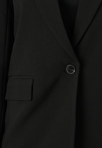 Musta bleiseri, jossa on lohkoläppä, yksittäinen napin kiinnitys ja pieni, viisto tasku. Kangas vaikuttaa sileältä ja istuu hyvin.