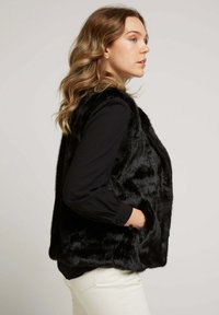 Gilet en fausse fourrure noire avec une texture lisse, des poches latérales et un ourlet arrondi. Porté par-dessus une chemise noire à manches longues, avec une coupe décontractée.