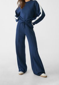 Completo di felpa e pantaloni in morbido tessuto blu navy. La felpa presenta strisce bianche sulle maniche; i pantaloni hanno una vita elastica e gambe ampie.