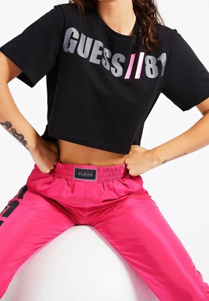 Osoba nosząca czarny krótki t-shirt z napisem „GUESS // 81” w szarym i różowym kolorze oraz jasnoróżowe spodnie z elastycznym pasem i metką marki.