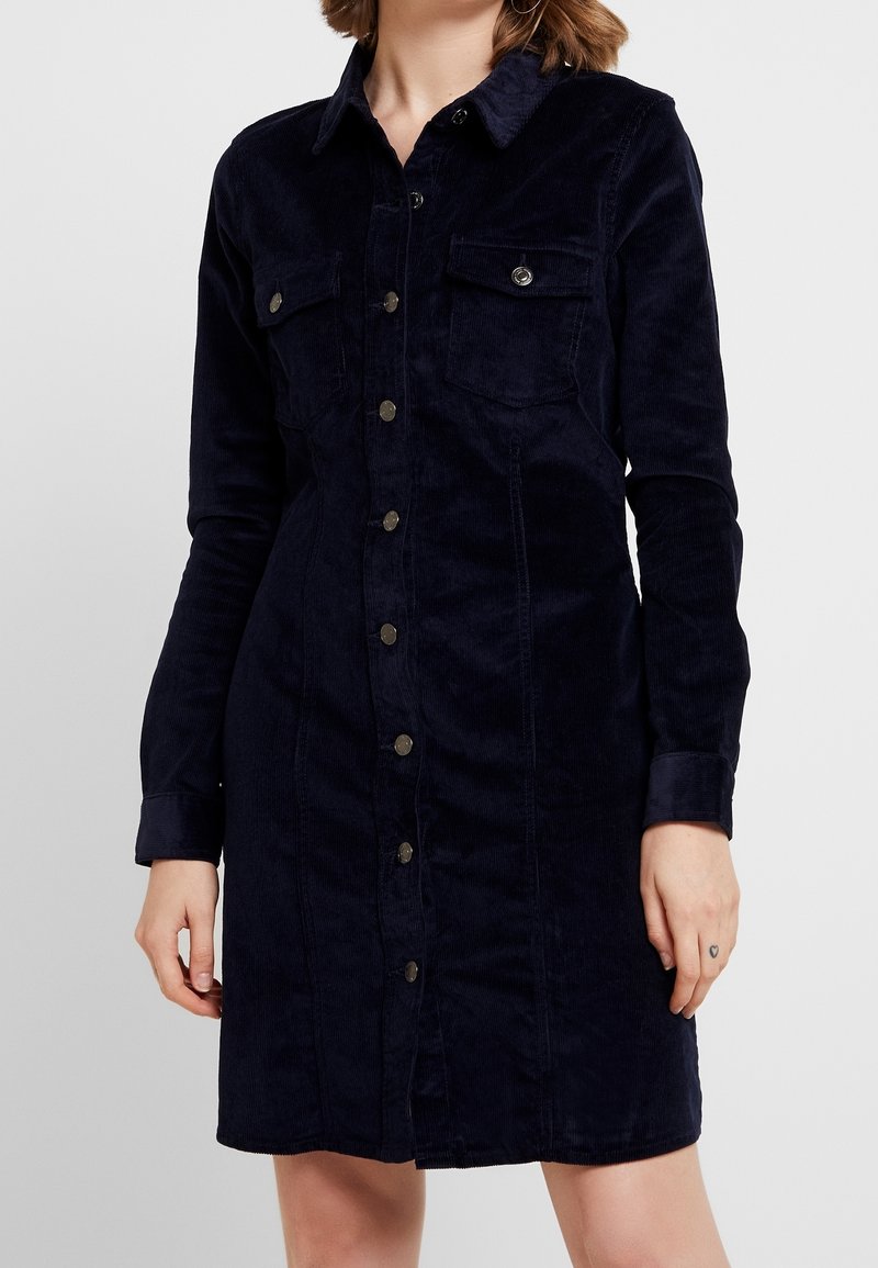 Femme portant une robe noire en velours côtelé boutonnée, manches longues et poches poitrine, debout devant un fond clair uni.