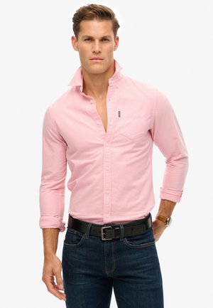 Lichtroze overhemd met knopjes, een kraag, lange mouwen en een borstzak. Draagt met donkerblauwe jeans en een zwarte riem. Eenvoudig, klassiek ontwerp.