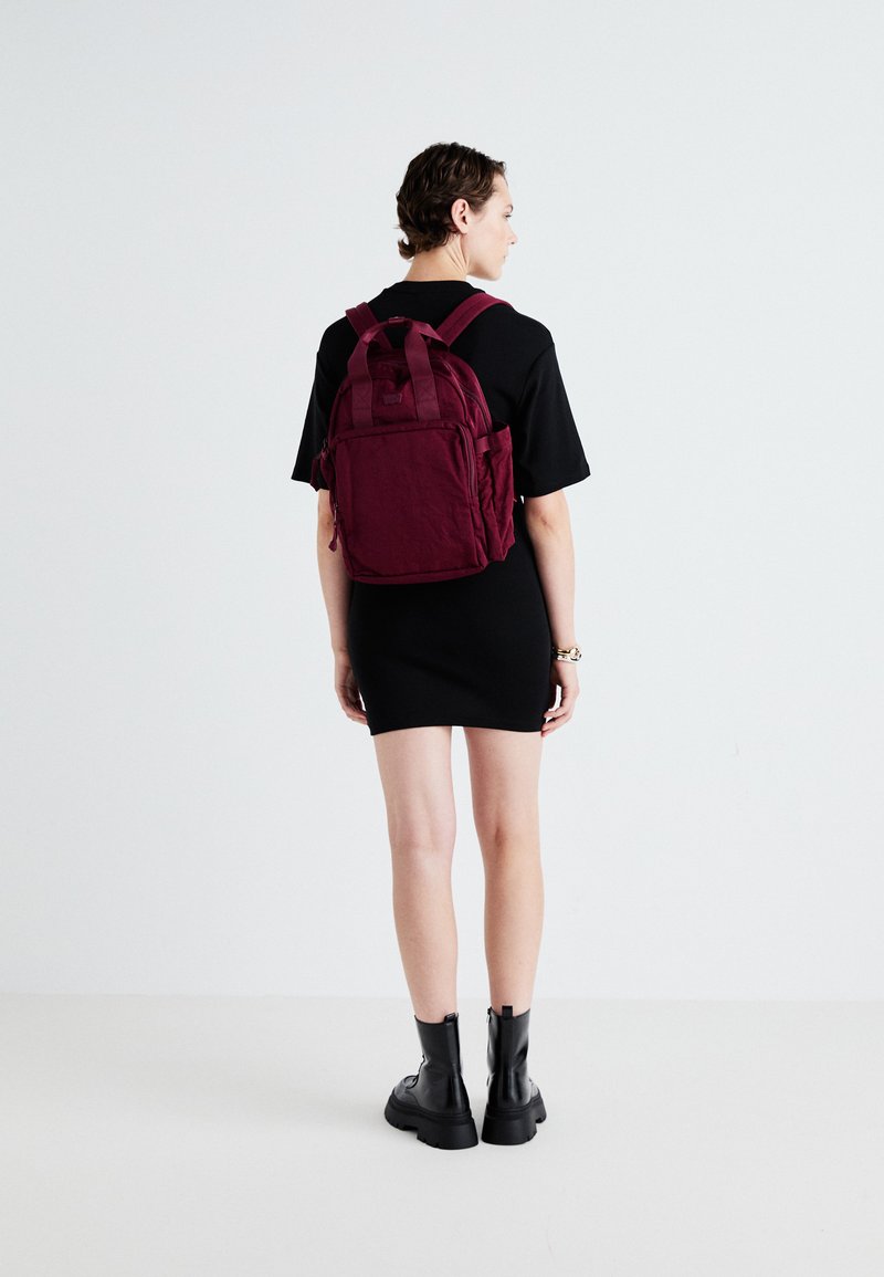 Bordeaux rygsæk lavet af tekstureret stof, med polstrede stropper, flere lommer og en kompakt, rektangulær form mod en minimal baggrund.