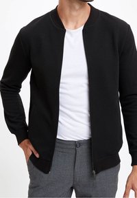 Czarna kurtka typu bomber zapinana na zamek, wykonana z teksturowanej tkaniny, z zaokrąglonym dekoltem, ściągaczami na rękawach oraz bocznymi kieszeniami. Noszona na białej koszulce i szarych spodniach.