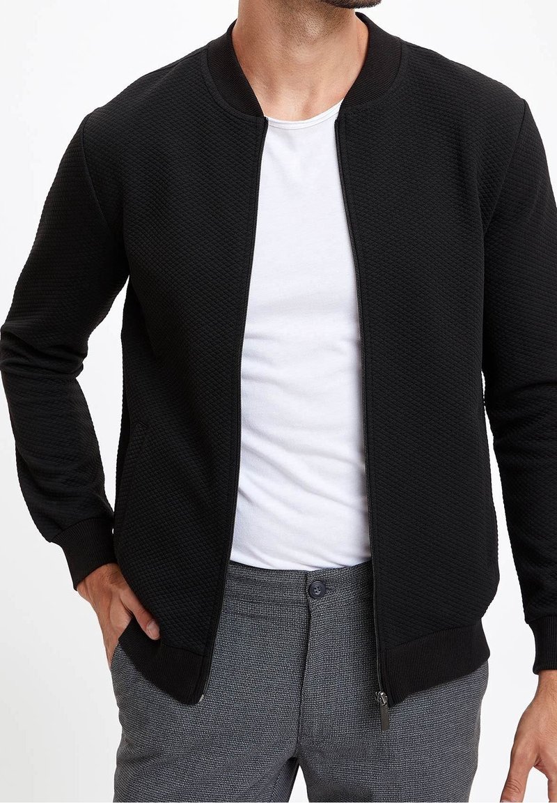 Czarna kurtka typu bomber zapinana na zamek, wykonana z teksturowanej tkaniny, z zaokrąglonym dekoltem, ściągaczami na rękawach oraz bocznymi kieszeniami. Noszona na białej koszulce i szarych spodniach.