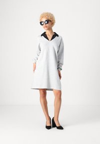 Vestido de sweat cinza com mangas longas, decote em V, colarinho preto em contraste, bolsos laterais e um silhueta descontraída, combinado com sapatos de salto pretos.