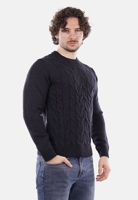 Uomo con capelli ricci che indossa un maglione lavorato a trecce blu navy e jeans blu, in piedi con una mano in tasca davanti a uno sfondo bianco.