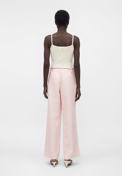 Γυναίκα που στέκεται με την πλάτη προς τα πίσω, φορώντας λευκή αμάνικη μπλούζα, ανοιχτόροζ παντελόνια με φαρδιά πόδια και λευκά παπούτσια με χαμηλό τακούνι, σε ένα απλό φόντο.