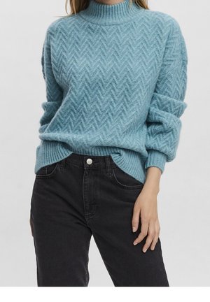 Femme portant un pull à chevrons bleu clair, manches retroussées, rentré dans un jean noir taille haute, sur un fond uni.