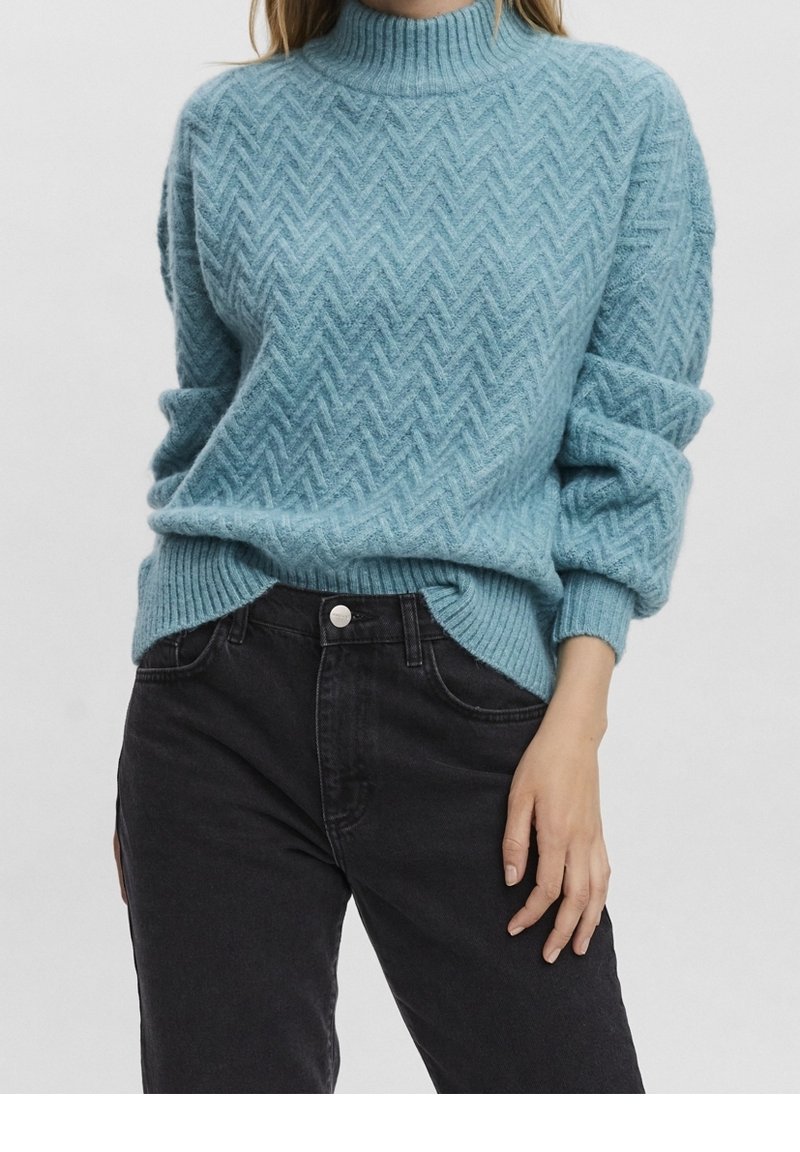 Femme portant un pull à chevrons bleu clair, manches retroussées, rentré dans un jean noir taille haute, sur un fond uni.
