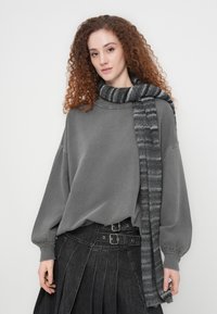 Γυναίκα με μακριά σγουρά κόκκινα μαλλιά που φορά χαλαρό γκρι φούτερ, ριγωτό πλεκτό κασκόλ και πλισέ μαύρη φούστα με διπλή ζώνη με αγκράφες.