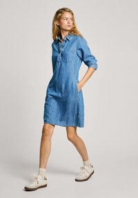 Denim srajčna obleka v svetlo modri barvi z dolgimi rokavi, gumbastim ovratnikom, žepom na prsih, stranskimi žepi in sproščenim krojem. V kombinaciji z belimi supergami.