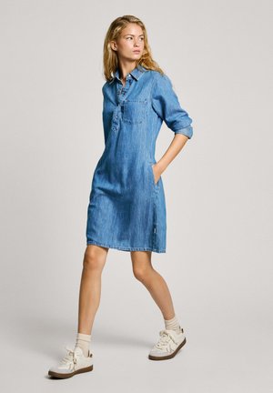 Vestido camisa de denim en azul claro con mangas largas, collar abotonado, bolsillo en el pecho, bolsillos laterales y un corte holgado. Combinado con zapatillas blancas.