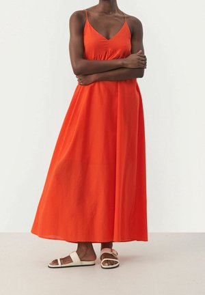 Kobieta w jasnopomarańczowej, bezrękawnikowej sukience maxi i białych, płaskich sandałach, stojąca z założonymi ramionami na tle jednolitego tła.