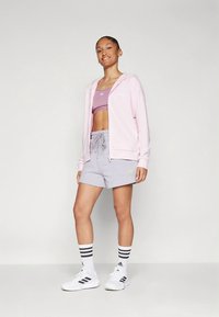 Gaiši rozā džemperis ar rāvējslēdzēju, valkāts virs lillā sporta krūštura un pelēkiem šortiem ar auklu. Baltas kurpes ar melnām svītrām un rakstainas zeķes papildina izskatu.