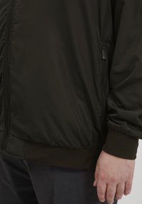Crna lagana bomber jakna s patentnim zatvaračem, rebrastim manžetama i glatkom, sjajnom teksturom. Vidljiva ruka koja odmara na rubu.