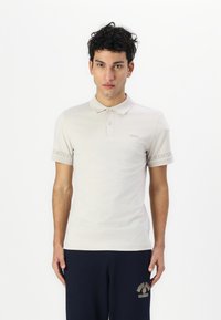Helles beiges Poloshirt aus Baumwolle, mit klassischem Kragen, kurzen Ärmeln und einem dezenten Logo-Detail am linken Arm.