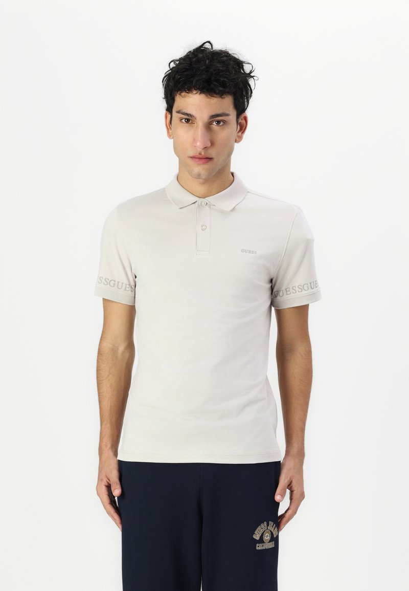 Helles beiges Poloshirt aus Baumwolle, mit klassischem Kragen, kurzen Ärmeln und einem dezenten Logo-Detail am linken Arm.