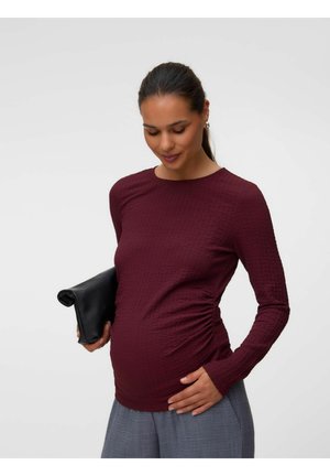 Femme enceinte portant un haut bordeaux texturé à manches longues, tenant une pochette noire et posant la main sur son ventre devant un fond uni.